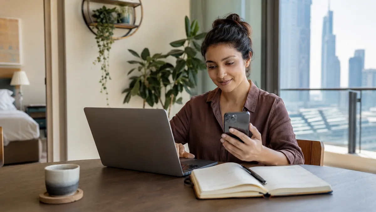A young woman in her Dubai apartment paying the rent on her phone, with a laptop and a planner on the table