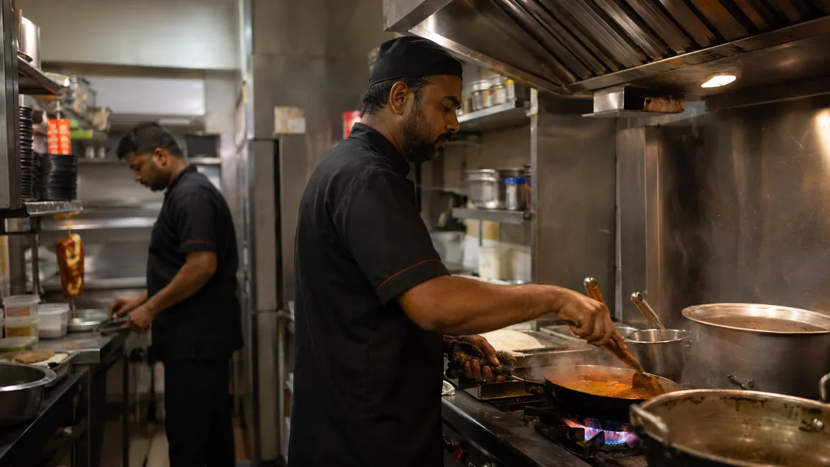 A chef and a helper working in a small Dubai restaurant kitchen with a steel exhaust hood and gas burners